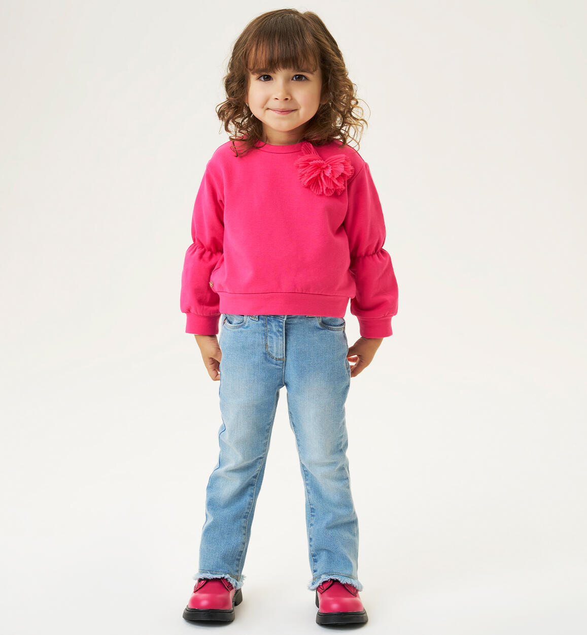 A girl wearing blue jeans and a pink sweater with a floral design on the chest, paired with pink shoes.