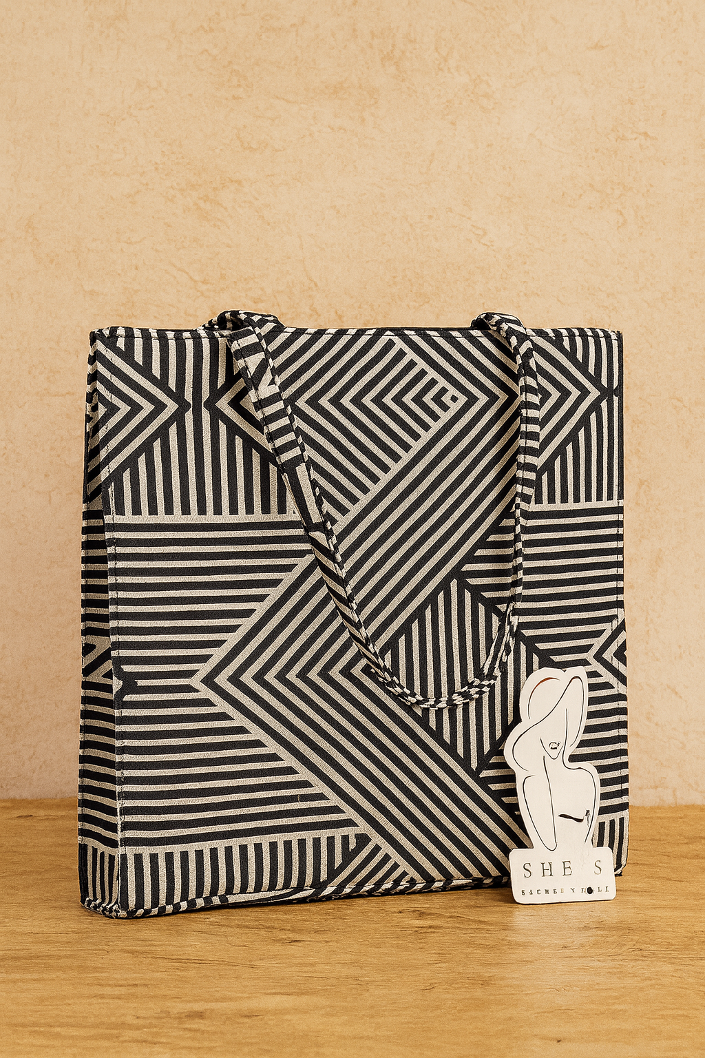 Black and white geometric patterned tote bag on a wooden surface with a small card labeled 'SHE'S' in the corner.