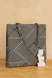 Black and white geometric patterned tote bag on a wooden surface with a small card labeled 'SHE'S' in the corner.