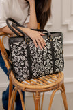 Black floral-patterned handbag held by a person on a wooden chair.