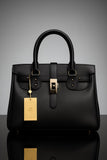 Black handbag with gold tag on a reflective surface and gray background