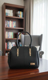 Black handbag with gold accents on a wooden table in a room with bookshelves and a chair.