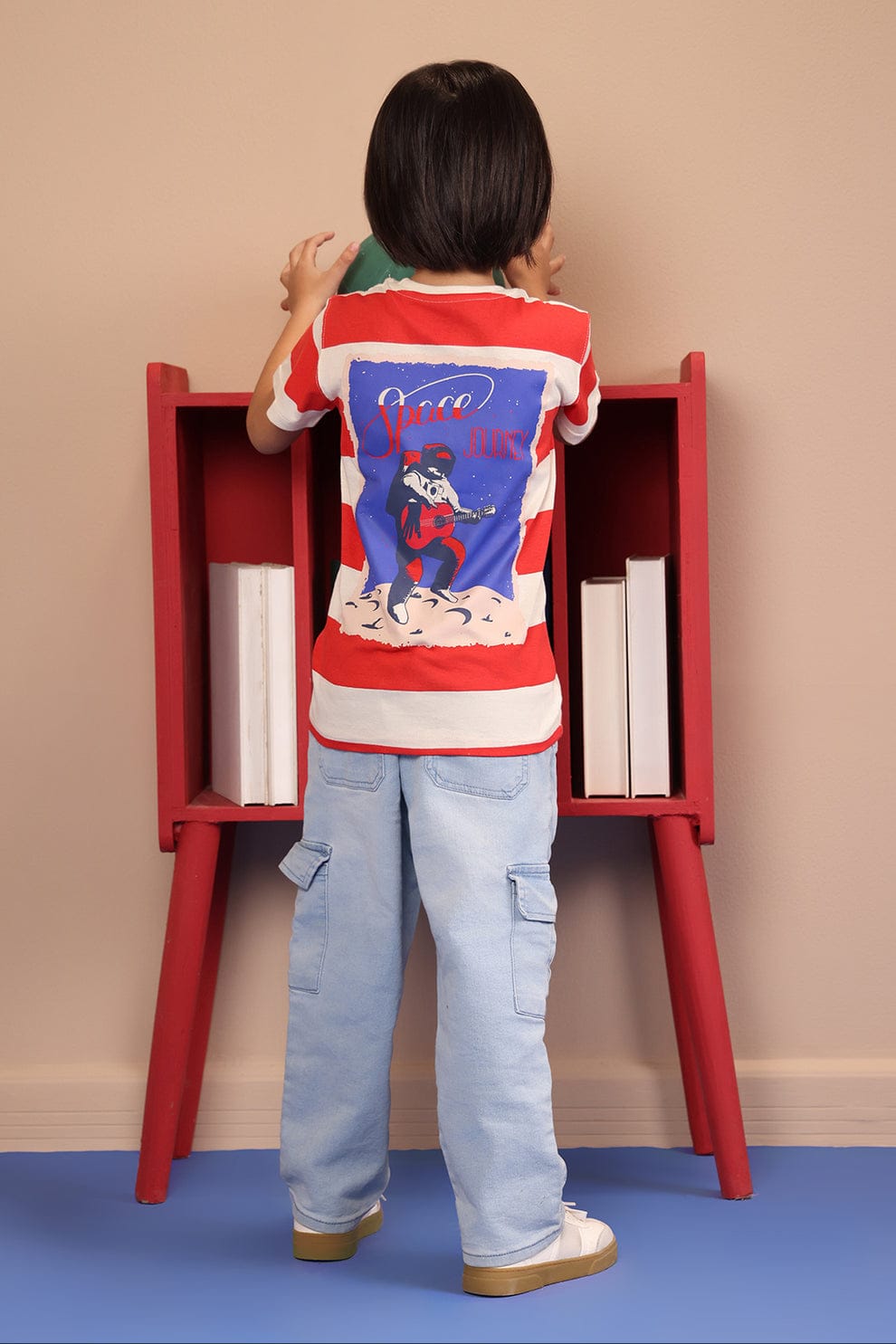 Child in a red and white striped shirt and light blue jeans standing next to a red bookshelf.