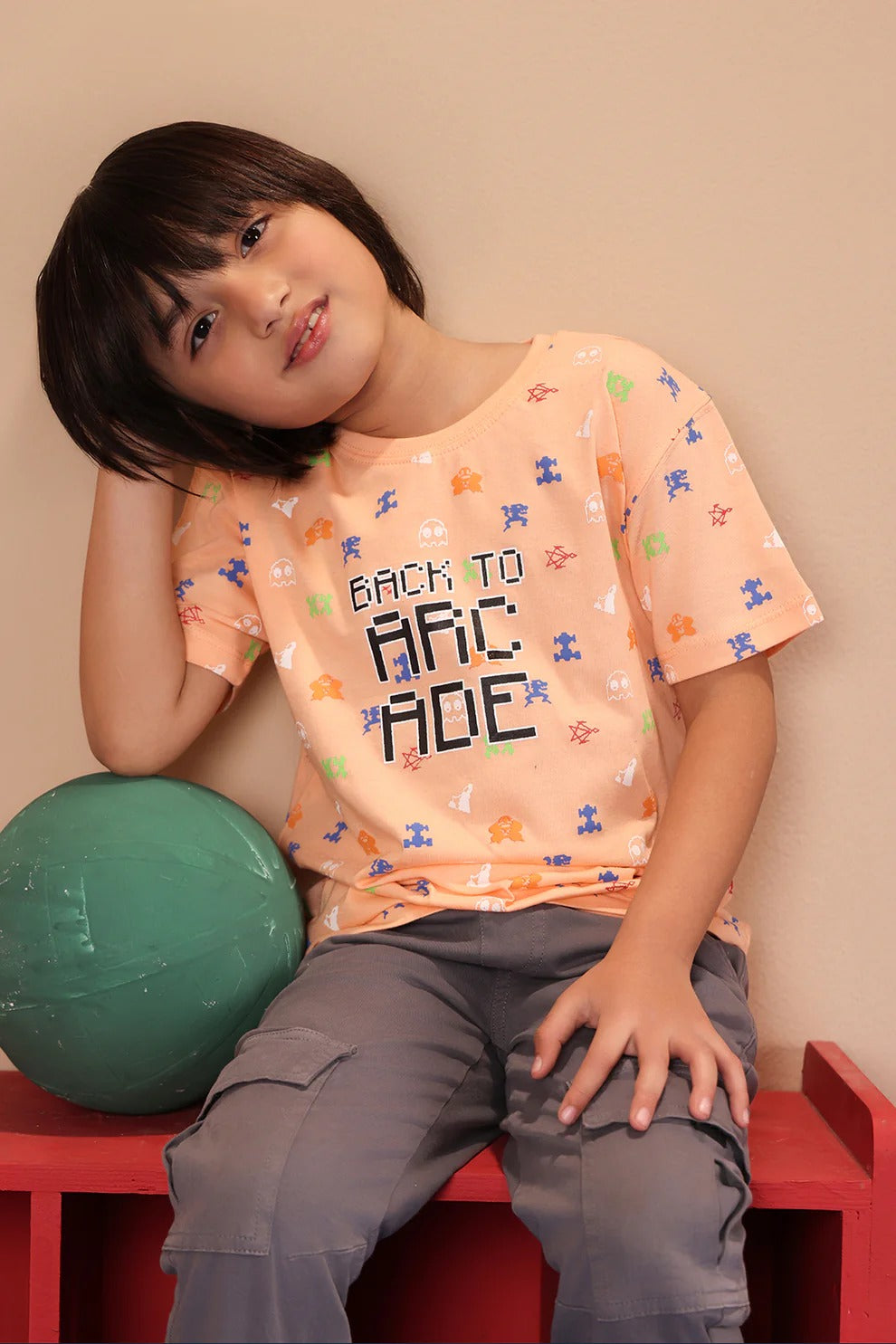 Child wearing a 'Back to Arcade' t-shirt, sitting on a red bench with a green ball.
