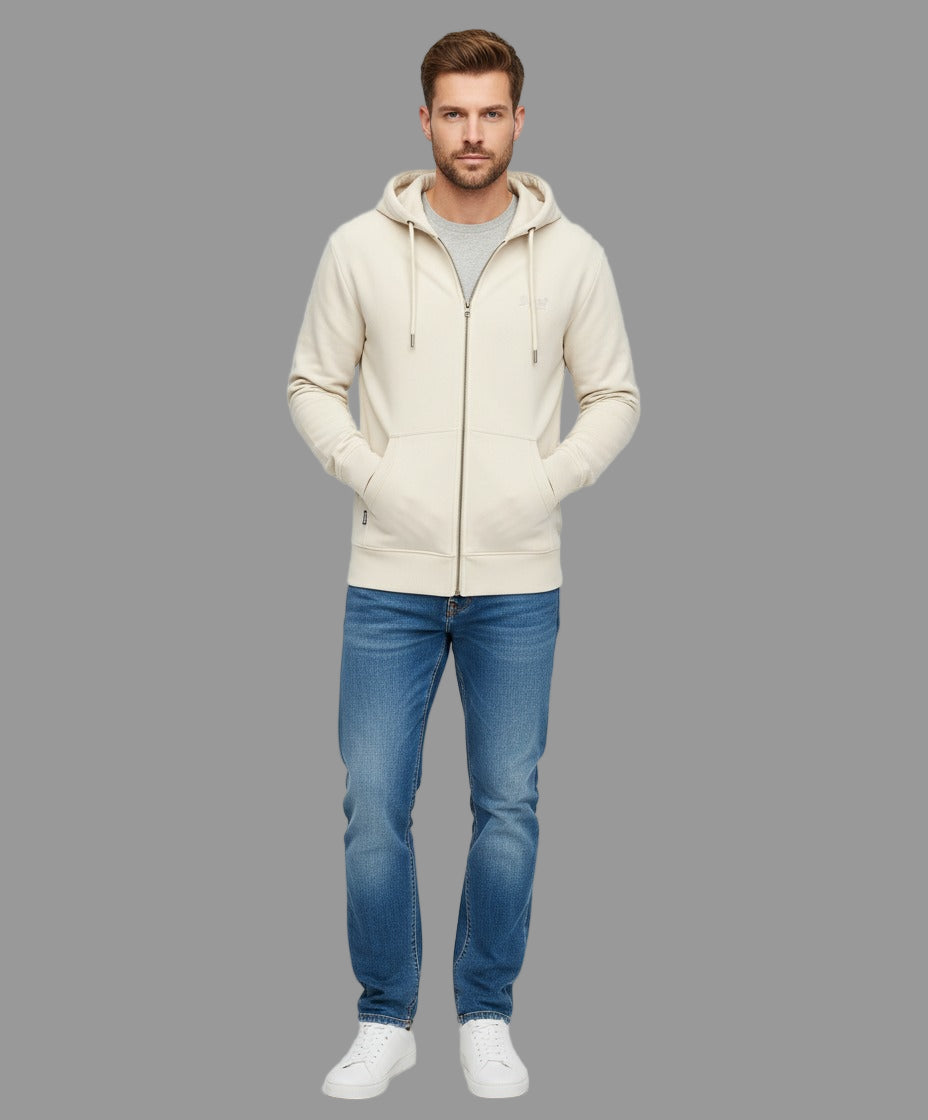 Man wearing a cream hoodie, gray shirt, blue jeans, and white sneakers on a light gray background