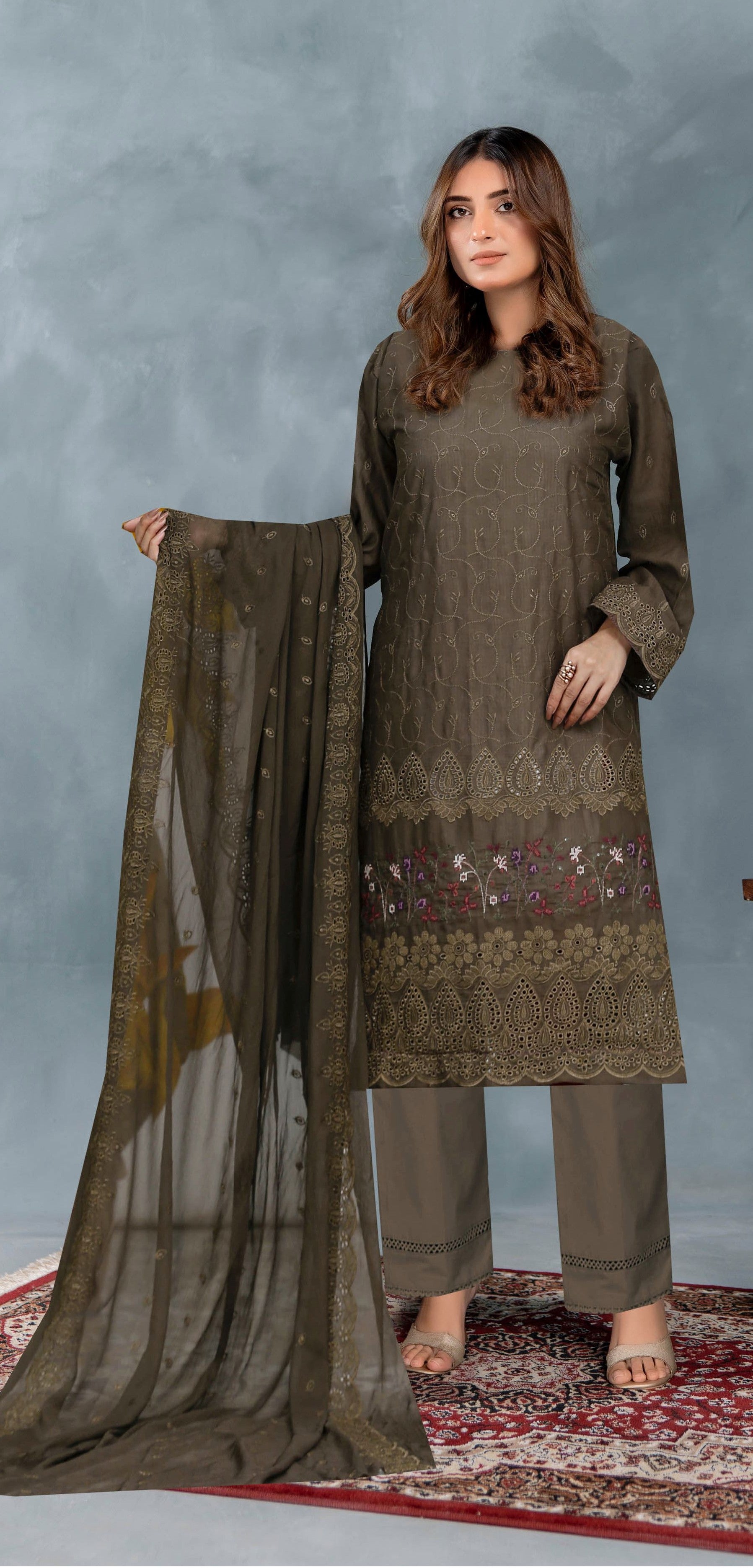 Woman holding a long scarf wearing a brown embroidered outfit on a gray background