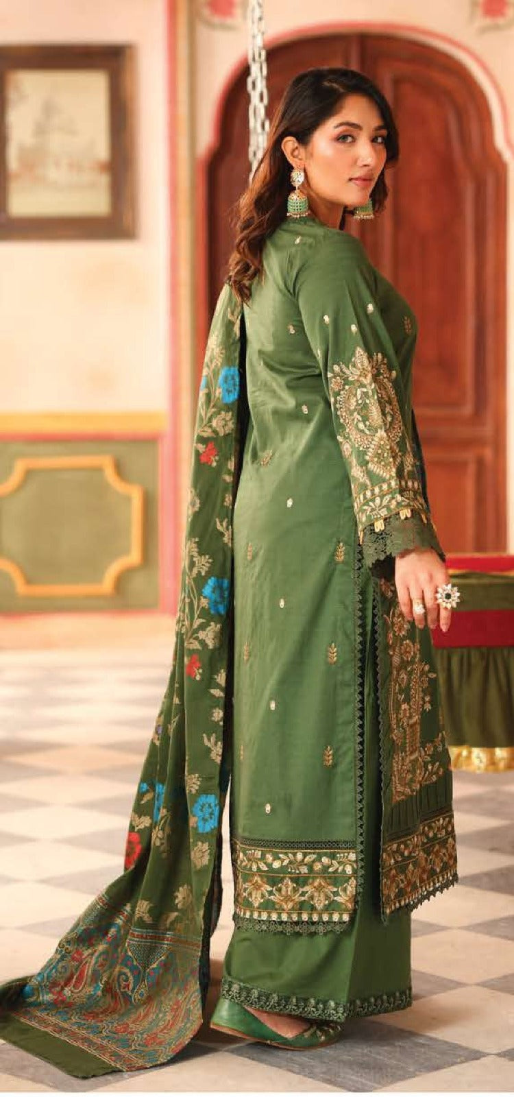 Woman in a green embroidered traditional outfit standing in a decorated interior setting.