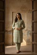 Woman in a light green traditional outfit standing in a sunlit room with textured walls.
