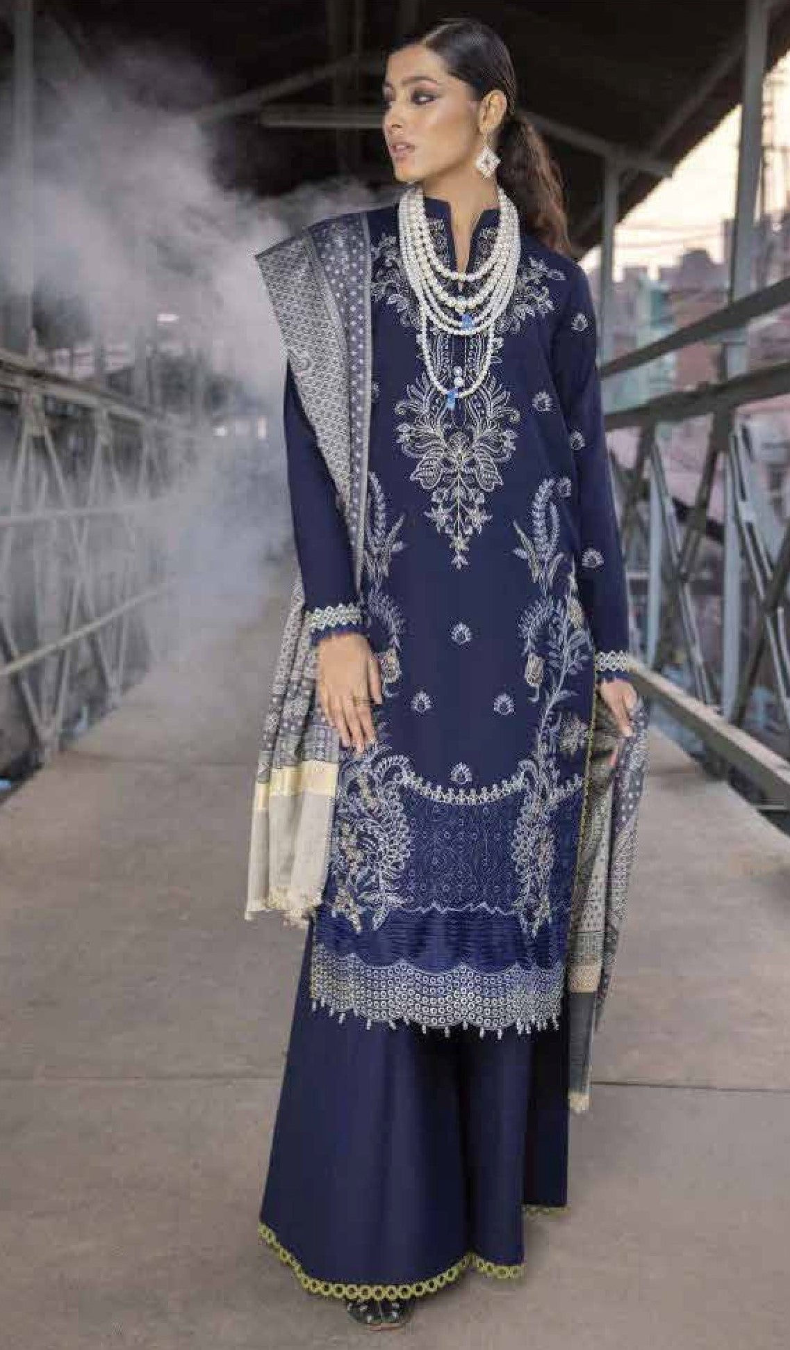 Woman in a navy blue embroidered traditional outfit with a dupatta, standing on a bridge with smoke in the background.