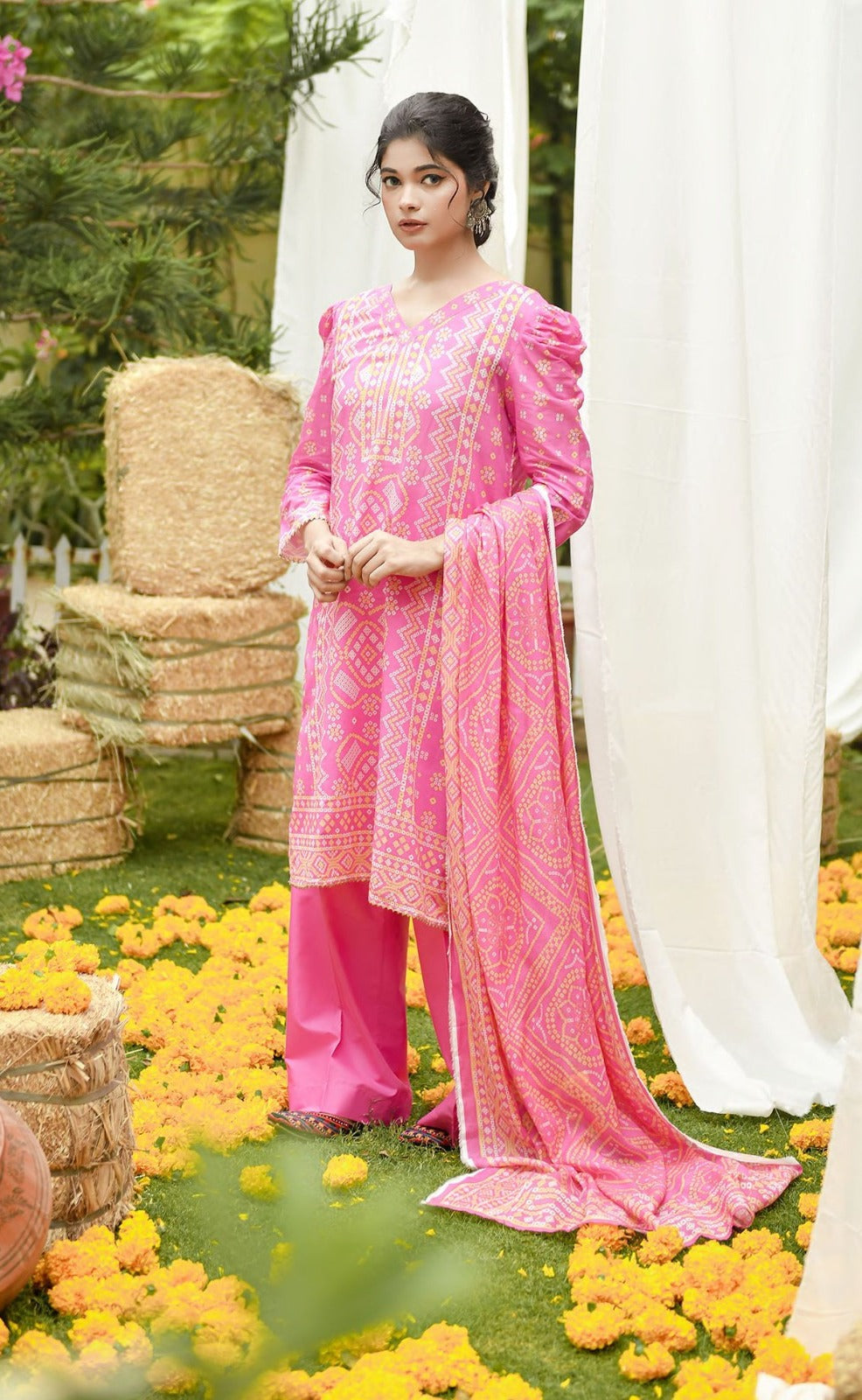 Woman in a pink traditional outfit standing outdoors with flowers and straw bales in the background
