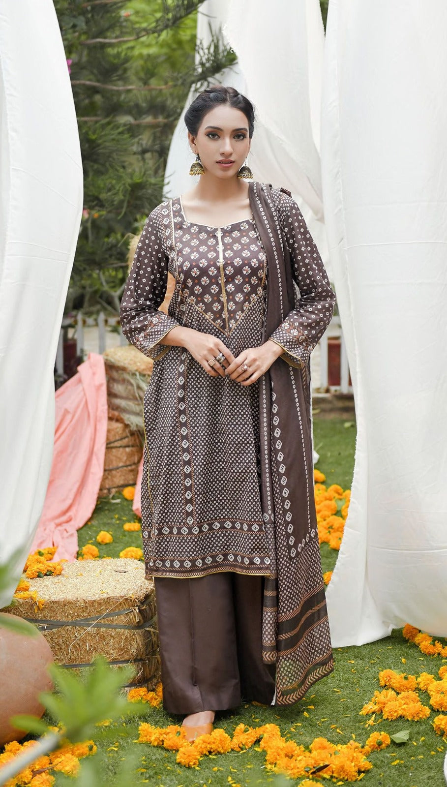 Woman in a traditional outfit standing in a decorated outdoor setting with flowers and white curtains.