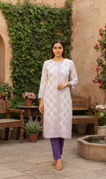 Woman in a white patterned kurta with purple pants standing in an outdoor setting with greenery and a fountain.