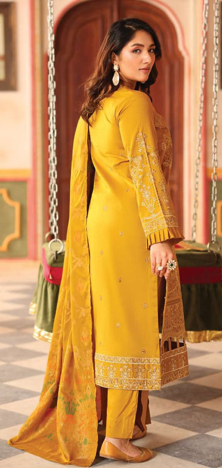 Woman in a yellow traditional outfit with intricate patterns, standing in a decorative indoor setting.