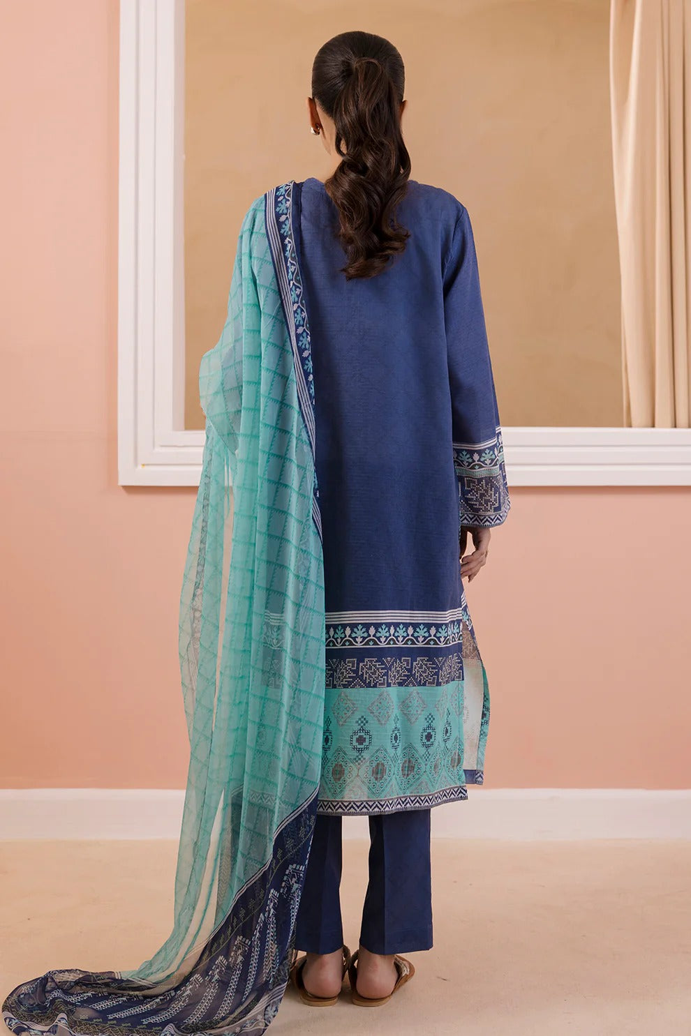 Woman wearing a blue outfit with a aqua green dupatta in a room with peach walls and a white frame.