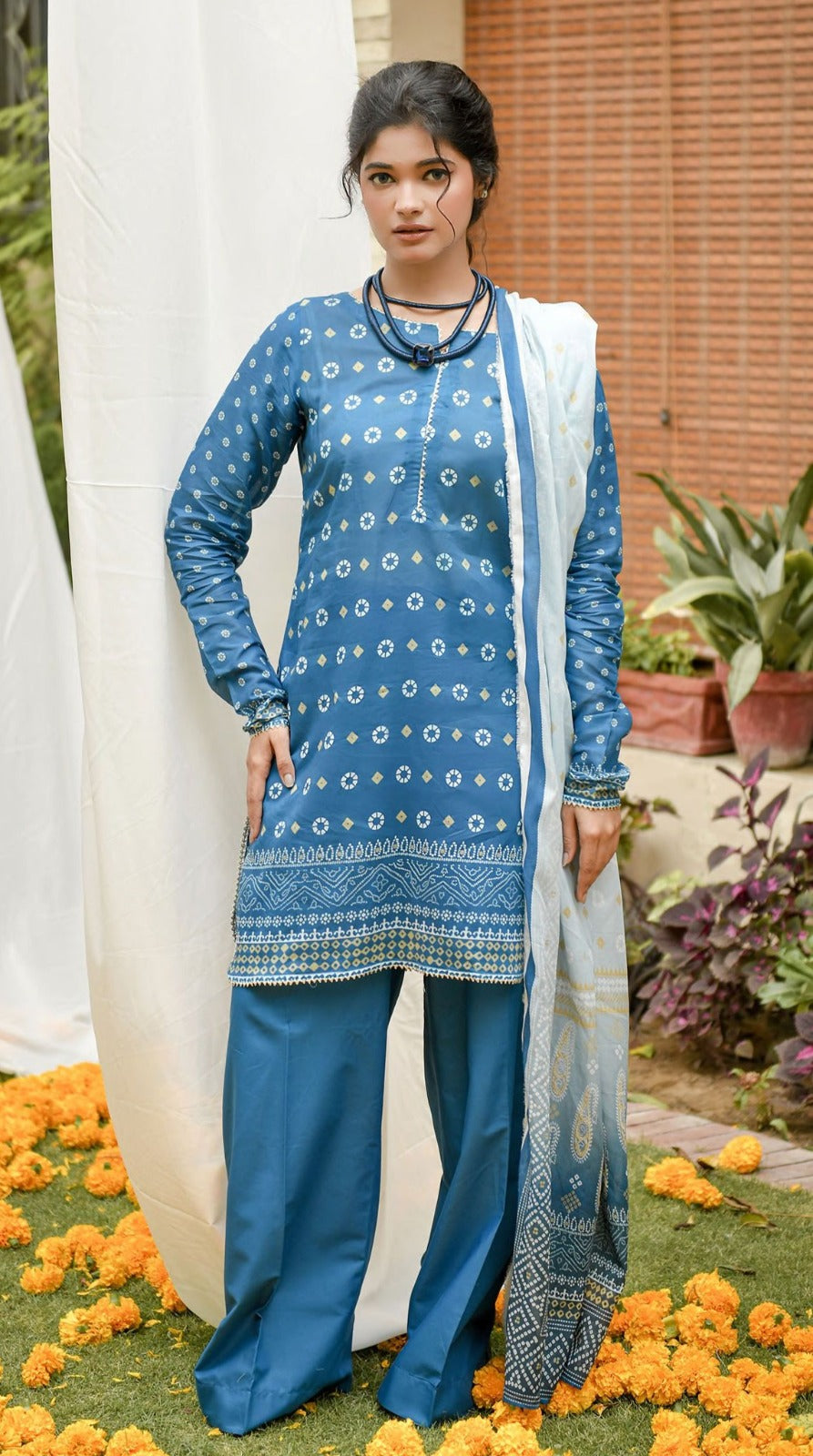 Woman wearing a blue patterned outfit with a white scarf, standing in a decorated outdoor setting.