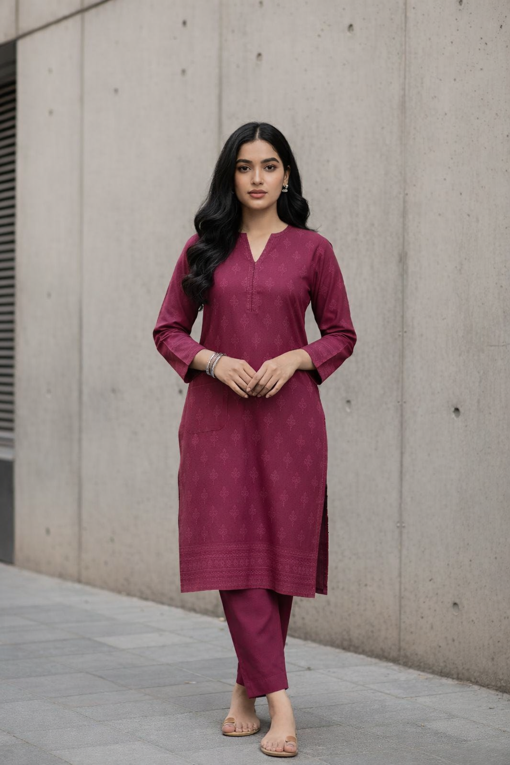 Woman wearing a darkpink kurta with pants standing against a concrete wall.