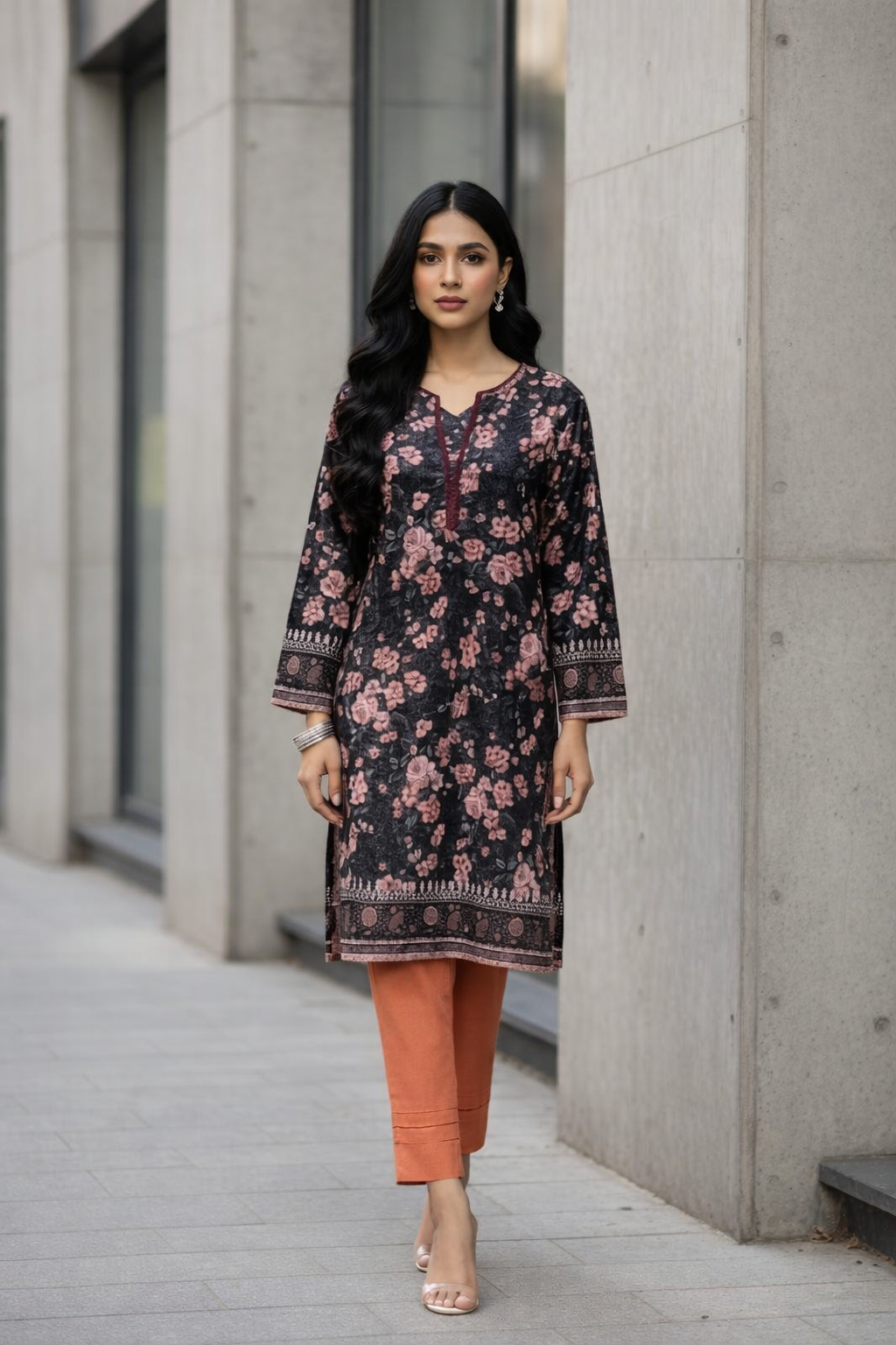 Woman wearing a floral dress and orange pants standing against a concrete wall.