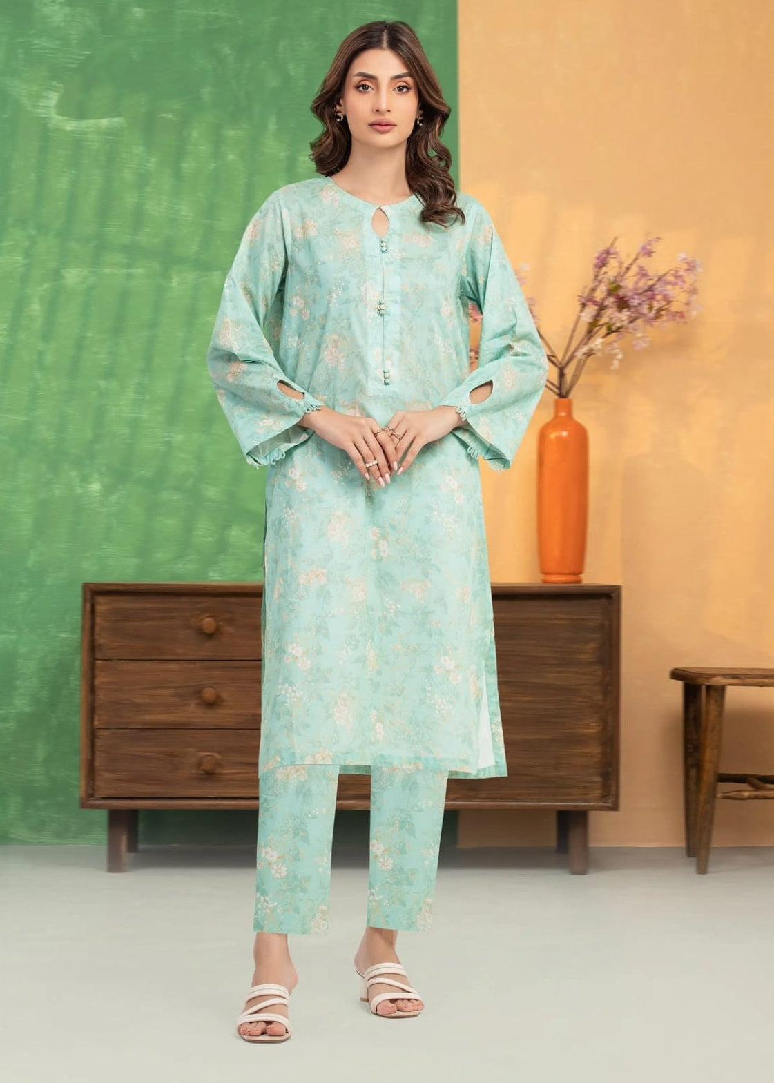 Woman wearing a light green floral outfit standing in a room with a green wall and wooden furniture.