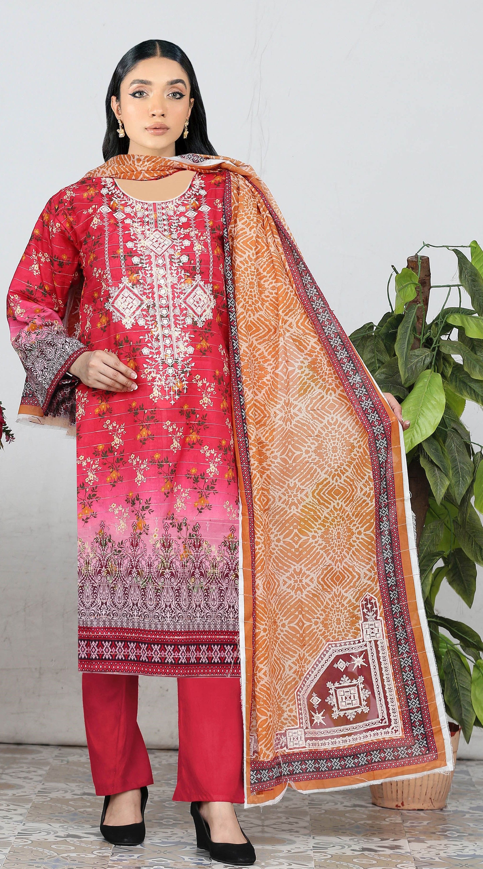 Woman wearing a red and pink embroidered outfit with a matching dupatta, standing against a white wall with a plant in the background.