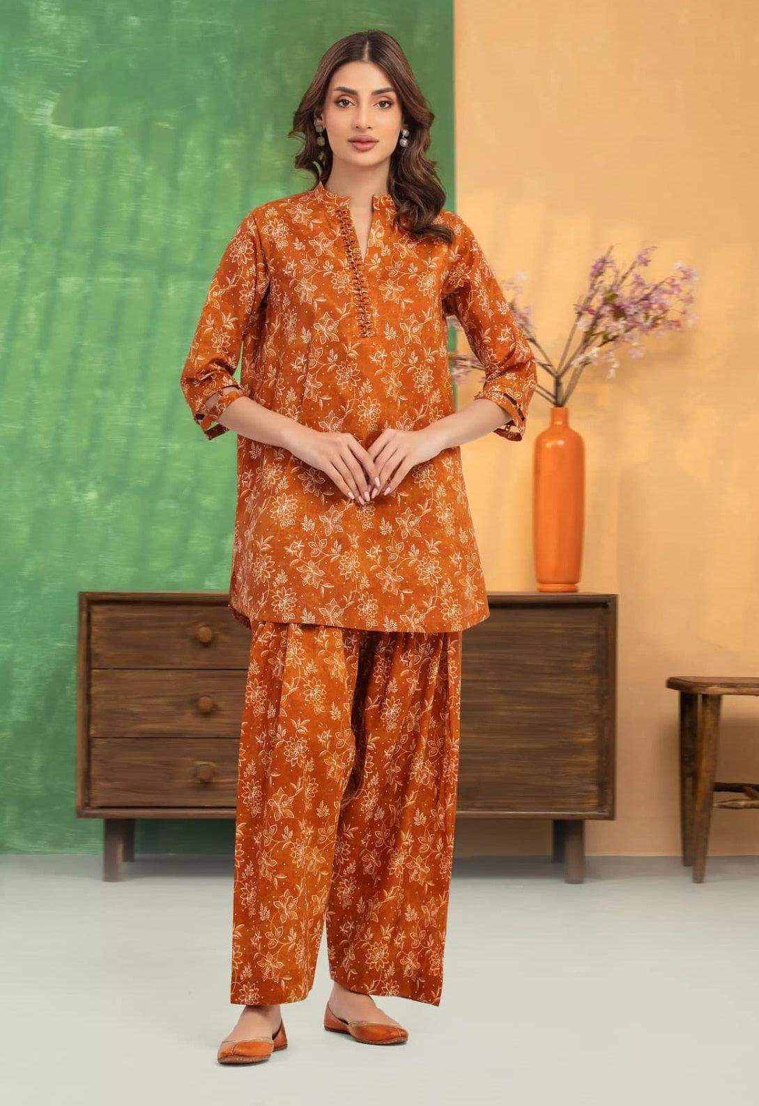 Woman wearing an orange floral outfit standing in a room with a green wall and wooden furniture.