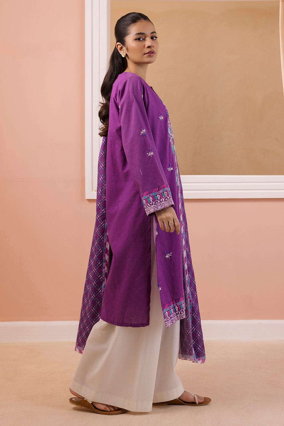 Woman wearing a purple embroidered traditional outfit against a beige wall.