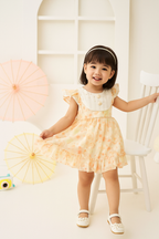 Young girl in a floral dress holding a orange parasol indoors.