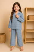 Young girl wearing a blue checkered outfit standing in front of a beige wall with a wooden shelf.