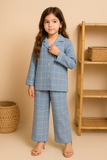 Young girl wearing a blue checkered outfit standing in front of a beige wall with a wooden shelf.