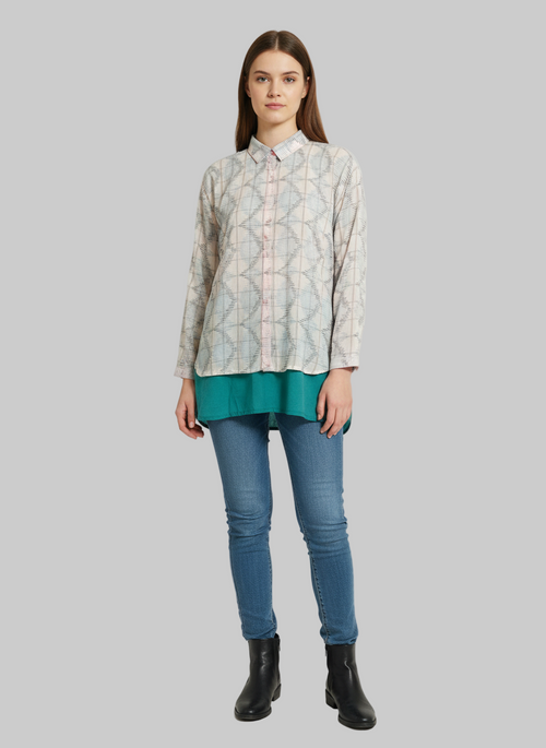 Woman wearing a patterned shirt, teal top, blue jeans, and black boots on a gray background