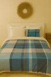Bed with plaid comforter and decorative pillow against a beige wall with a decorative wreath.
