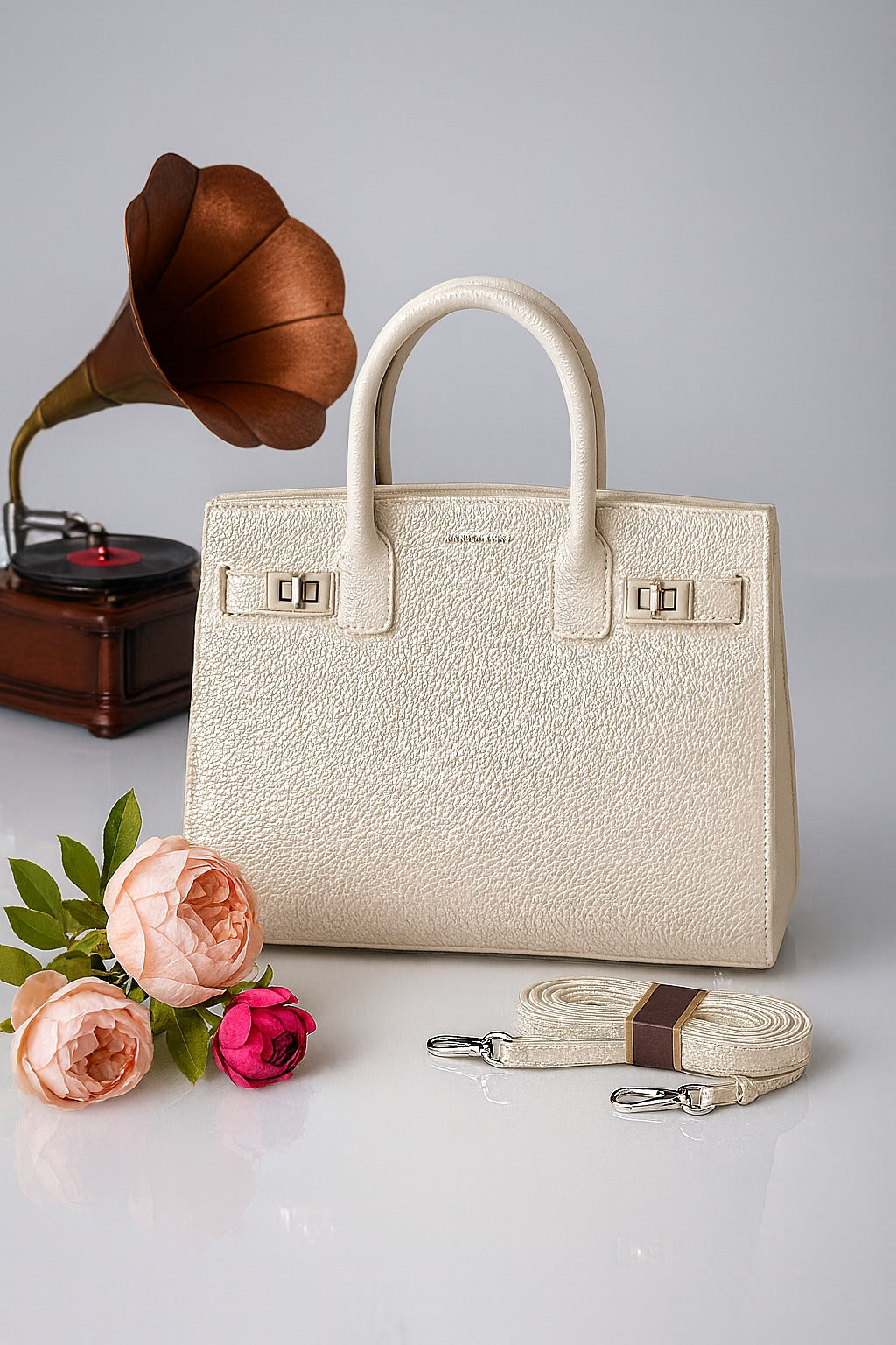 white handbag with a matching strap on a light gray background, accompanied by flowers and an old-fashioned record player.