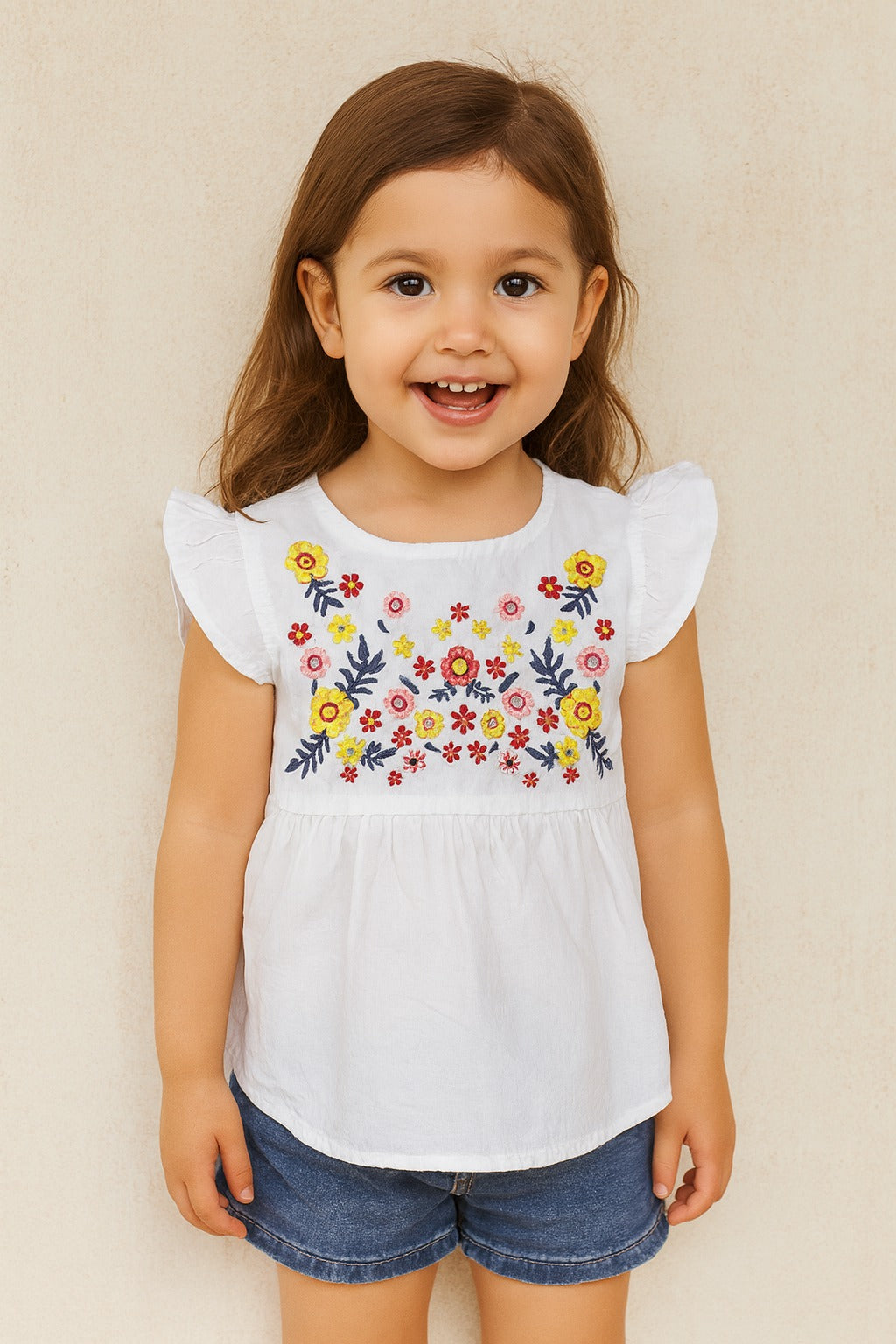 Young girl wearing a white top with floral embroidery and denim shorts against a beige background