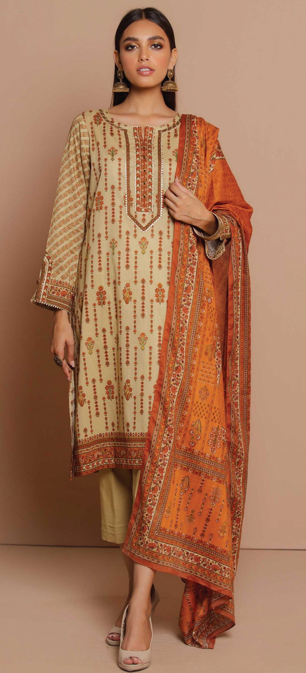 Woman wearing a traditional outfit with a beige kurta, orange dupatta, and green pants against a beige background.